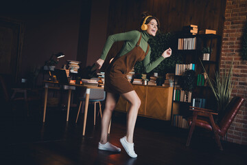 Naklejka premium Young woman in brown jumper dress joyfully dances in her cozy apartment while studying, surrounded by books