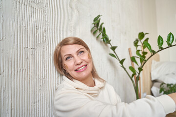 Smiling caucasian adult female sitting near textured wall with green plant decor.