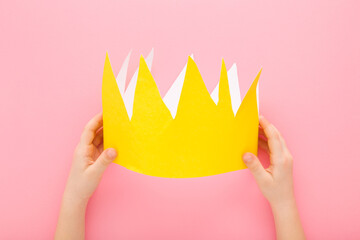 Little child girl hands holding and showing yellow crown from application paper on light pink table...