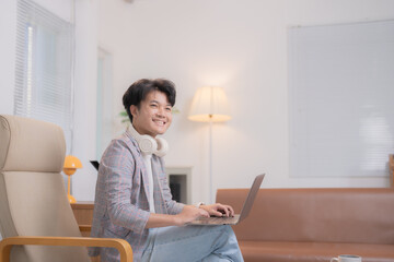 Young asian businessman is sitting comfortably in a chair in his modern living room, working on his laptop with a smile and headphones around his neck, enjoying the benefits of remote work