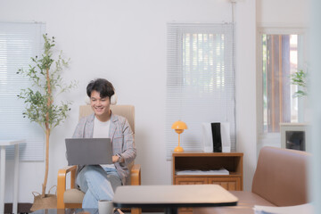 Young asian man is working from home, sitting comfortably in an armchair, using laptop and headphones, enjoying the convenience and flexibility of remote work in a bright and cozy home office