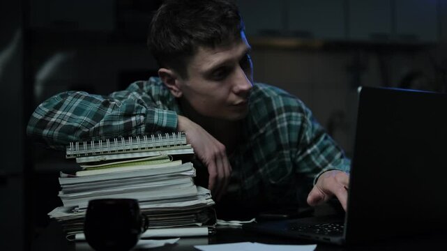 Student while studying for exams fell asleep at night on desk with cup of coffee. Wakes up and continues working on a laptop surrounded by notes. Fatigue and deadlines overwhelm him.