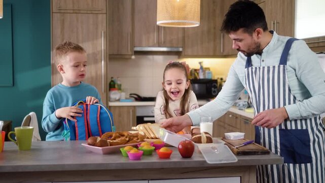 A young father and two small children make lunch box sandwiches together in the kitchen at home before going to kindergarten.