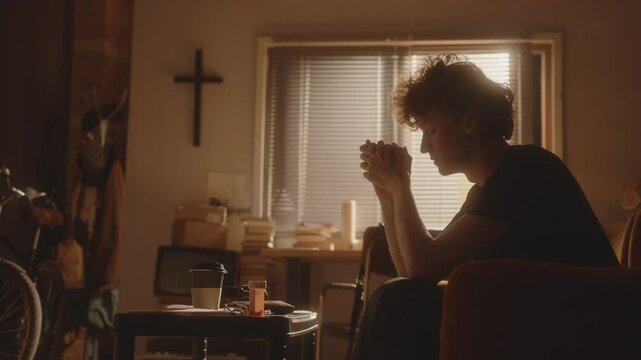 Vulnerable young man, full of sadness, sitting in armchair with hands clasped and praying to God, with cross on wall behind him and prescription bottle on table in softly lit living room