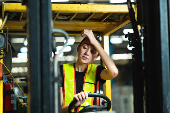 A woman in a yellow vest is driving a forklift. She is sweating and she is in pain