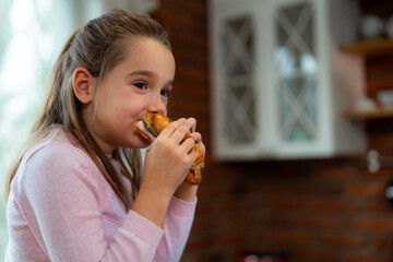 Caucasian girl with long brown hair, wearing a pink sweater, savoring a croissant sandwich filled...