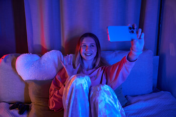 A cheerful teenage girl relaxes in her cozy room, capturing the moment on her phone.