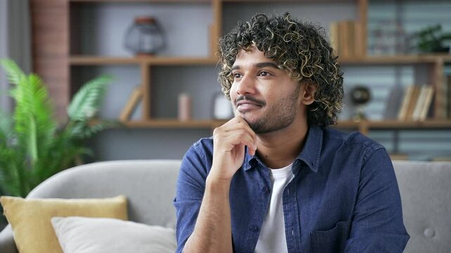 Thoughtful happy young man sitting on sofa in living room at home. Handsome male thinking about positive, daydreaming. Calm male looking away enjoying single life leisure time at weekend, relaxing