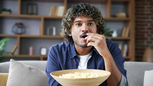 Happy man watching an interesting movie on television sitting on sofa in living room at home. Smiling male eating popcorn and watching show or TV series spending his leisure time. Front view. Close up