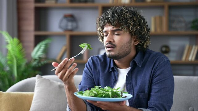 Tired of dieting for weight loss handsome man with a plate of salad sitting on the sofa in living room at home. Dissatisfied young male holds a fork in his hand, but does not want to eat healthy food