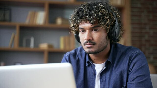 Focused IT specialist in wireless headphones works remotely on laptop sitting in living room at home office. Serious handsome coder developer is programming, busy with a project on computer. Close up