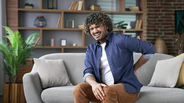 Tired young adult man suffering from back pain sitting on sofa in living room at home. Upset handsome male holds his lower back with his hands, massages painful muscles, has sciatica or a sick spine