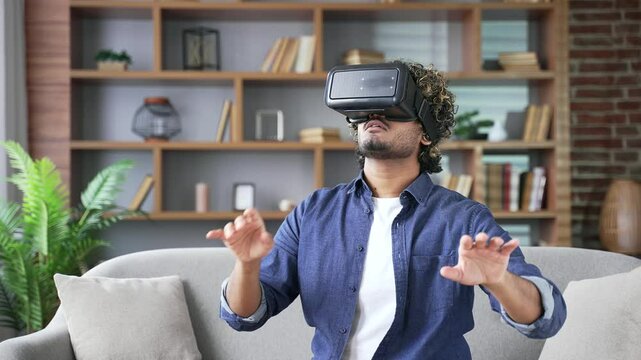 Confident young man works using VR glasses in virtual reality simulator sitting on sofa in living room at home office. Male uses gestures to control computer programs and flips through virtual pages