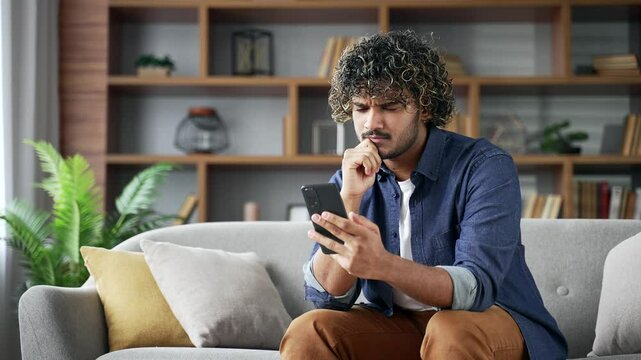 Thoughtful young man holding mobile phone sitting on sofa in living room, looking at screen with focus and curiosity. Serious handsome man using smartphone, reading message and thinking at home