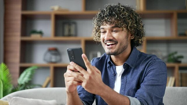 Satisfied happy young man received great news on mobile phone while sitting on sofa in living room at home. Smiling glad handsome male reads a positive good message on a smartphone, rejoices. Close up
