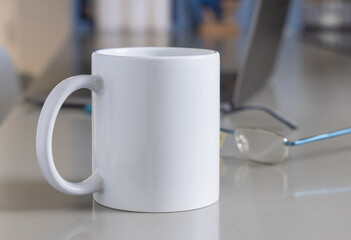 White coffee mug on grey office table against opened laptop and glasses closeup, mockup
