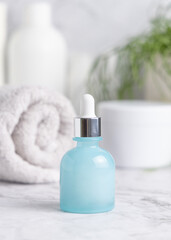 Light blue dropper bottle on marble near cosmetics, towel and green plant close up, mockup