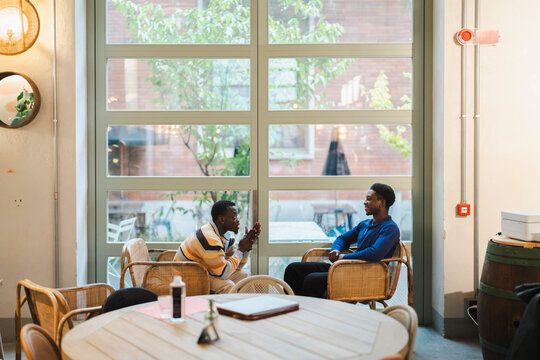 Two black men sitting in a modern cafe environment, deeply engaged in a meaningful conversation while seated in wicker chairs by large windows with natural light.