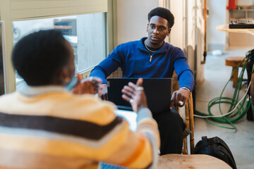 A young black man seated on a wicker chair with a laptop, appearing thoughtful and reflective, in a brightly lit cafe with a modern and inviting atmosphere enhanced by large glass windows.