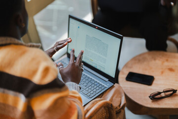 A young black man actively gesturing with his hands while explaining a concept on his laptop during a meeting in a cozy cafe environment with wooden furniture and warm lighting.