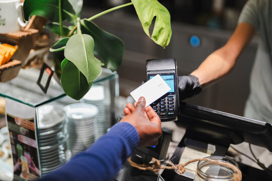 A hand holding a credit card extends towards a payment terminal in a modern cafe, surrounded by stylish decor and a glass display, showcasing a seamless and contemporary payment experience.