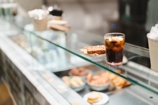 A glass counter in a cafe displays a slice of cake and a glass of iced drink alongside other baked goods, offering a glimpse of the appealing and carefully curated menu items in a modern setting.