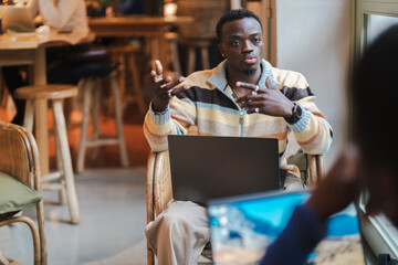 A black man seated in a wicker chair with a laptop gestures expressively while speaking, creating a dynamic and interactive moment in a cozy and modern cafe setting with warm tones and casual decor.