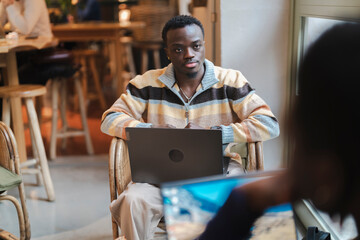 A black man seated in a wicker chair with a laptop gestures expressively while speaking, creating a dynamic and interactive moment in a cozy and modern cafe setting with warm tones and casual decor.