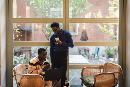 A black man standing with a latte in hand observes his friend working on a laptop in a bright cafe with large windows, natural lighting, and a calm and collaborative atmosphere.