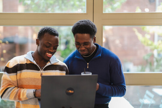 Two black friends in a cafe look at a laptop together near large windows with natural light and greenery outside, enjoying a collaborative and engaging moment in a modern and cozy environment.
