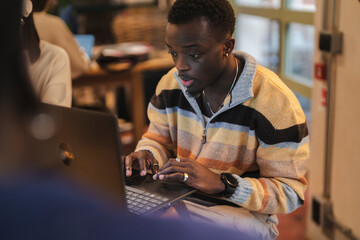 A young black man concentrates on his laptop during a collaborative session in a cozy cafe, where technology, creativity, and a relaxed atmosphere intersect seamlessly in a modern environment.