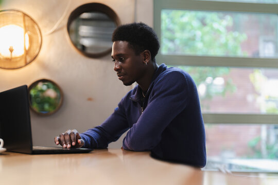 A young black man works on a laptop at a wooden table in a bright, modern workspace featuring natural lighting, greenery, and decor that encourages a calm and focused atmosphere for individual tasks.