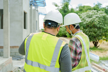 Engineer and architect collaborating at house construction site, holding tablet and blueprints to assess progress. Wearing safety helmets and reflective vests while discussing project execution.