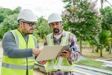 Engineer and architect analyzing house construction details at site with laptop and tablet. Wearing safety helmets and reflective vests, ensuring structural quality and project alignment.