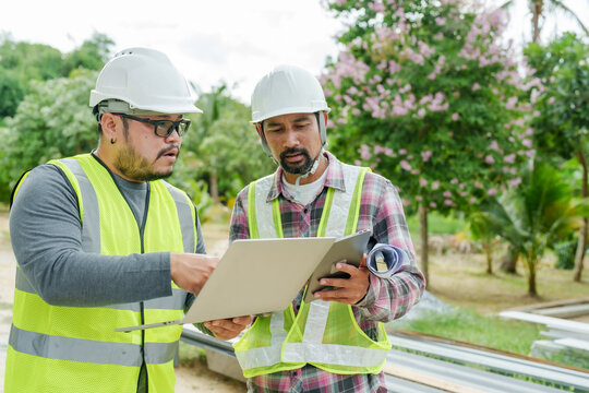 Engineer and architect analyzing house construction details at site with laptop and tablet. Wearing safety helmets and reflective vests, ensuring structural quality and project alignment.