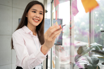 Young Asian woman working on project plan using sticky papers notes on glass wall for writing...