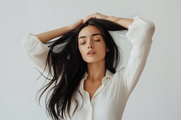 Naklejka premium Young woman with long black hair wearing a white shirt is pulling her hair with closed eyes, showing frustration on a white background