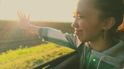 Traveling woman in car stretches hand out of car window, catching fair wind, rays of golden sun. Happy female traveler rides in car, enjoys road trip out of town, watches views of agricultural fields.