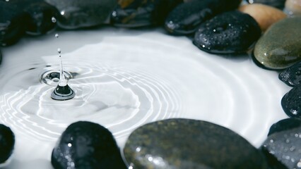 Water droplets create ripples in still water surrounded by smooth pebbles in a serene setting