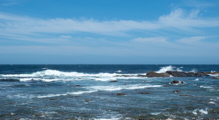 Coastal scene, ocean waves breaking on rocky shore, blue sky with clouds.