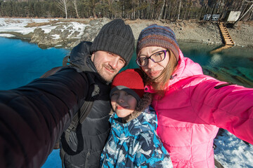 A family bundled in winter clothing taking a selfie by a scenic blue mountain lake surrounded by snow and forest.
