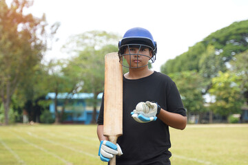 Cricket player wearing cricket Helmet holds a bat and ball in his hand, standing on the grass while...