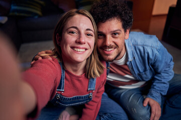 Caucasian couple at home taking a smiling selfie looking at camera to share on social media, having fun together in new apartment. POV people lifestyle.