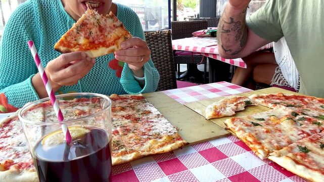 Tourists dining outdoors on a sunny terrace at an Italian restaurant, enjoying a Mediterranean meal with pizza and fresh ingredients, dressed casually in springtime or summer fashion.