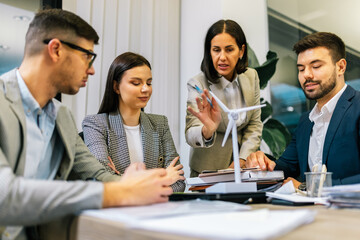 Bbusiness people talking about sustainable energy. They have windmill on a table and they are doing work on a laptop.