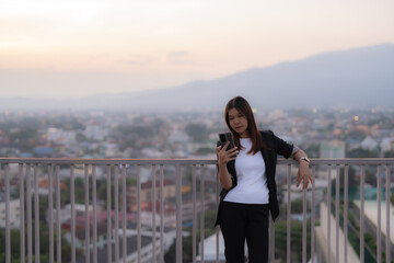 Young businesswoman is using her smartphone while leaning on a railing on a rooftop at sunset, overlooking a cityscape, enjoying the urban view