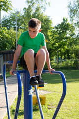 Fototapeta premium A focused young boy performs dips on outdoor fitness equipment. The setting is a sunny park with green grass and trees in the background. He wears a green shirt and dark shorts.