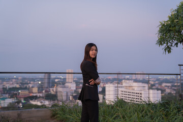 Fototapeta premium Young businesswoman standing on rooftop with arms crossed, enjoying cityscape view at dusk, showcasing confidence, ambition, and success in urban business environment