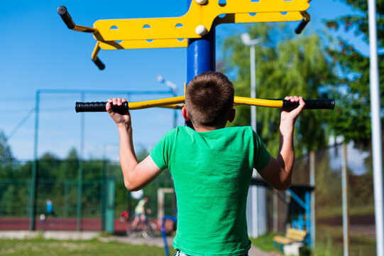 A young boy performs pull-ups on outdoor exercise equipment. The scene is set in a park on a bright sunny day, showcasing an active and healthy lifestyle.