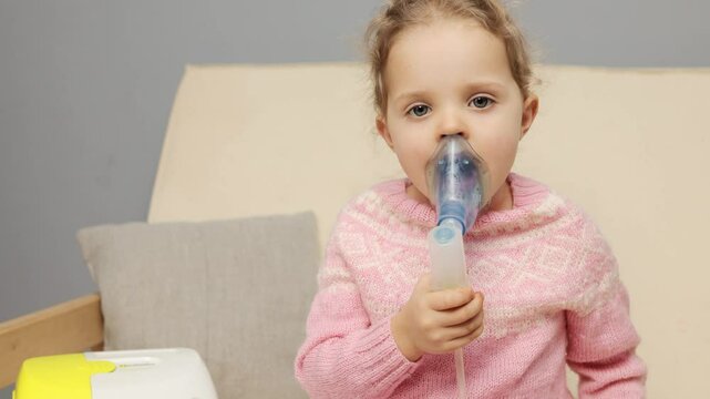 A young girl sits comfortably at home, inhaling medication through a nebulizer while wearing a cozy sweater. This treatment is crucial for her breathing health and well-being